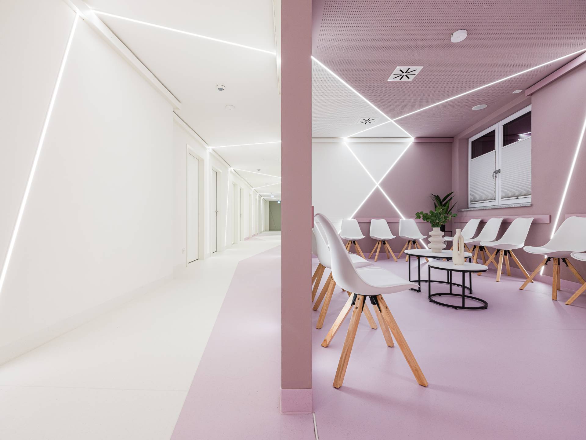 Moderner Flur mit weißer Wand, rosa Akzenten am Boden und geometrischem LED-Lichtdesign an der Decke und Wänden, der in einen Wartebereich übergeht mit weißen Stühlen mit Holzbeinen, einem schwarzen runden Tisch und dekorativen Pflanzen sowie einem Fenster mit Jalousien, aufgenommen von Architekturfotograf Alexander Gerner für das Augenzentrum Cham, gestaltet von Planquelle Straubing.