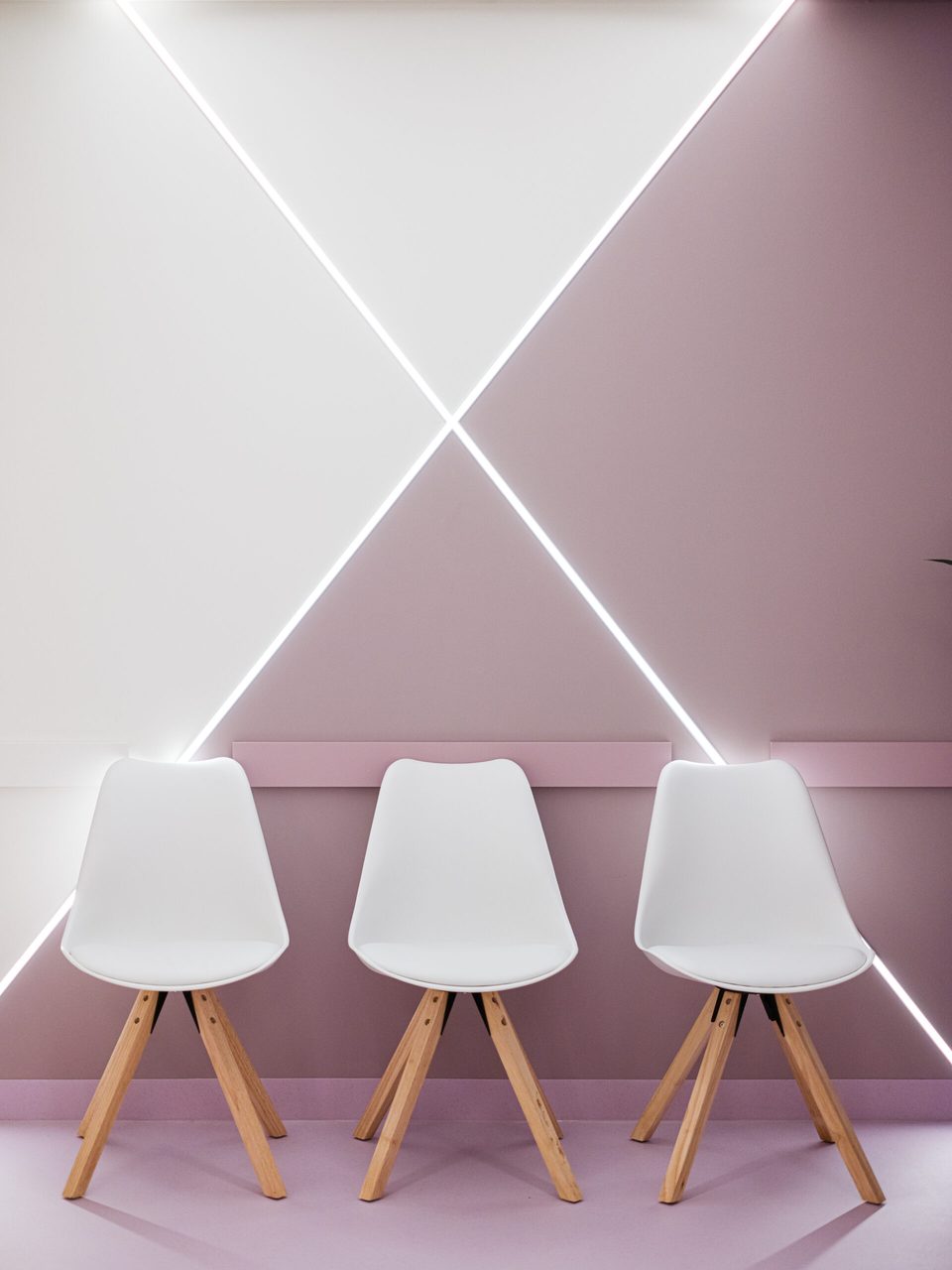 Drei weiße Schalenstühle vor roséfarbener Wand, über ihnen kreuzen sich zwei LED-Lichtlinien zu einem X