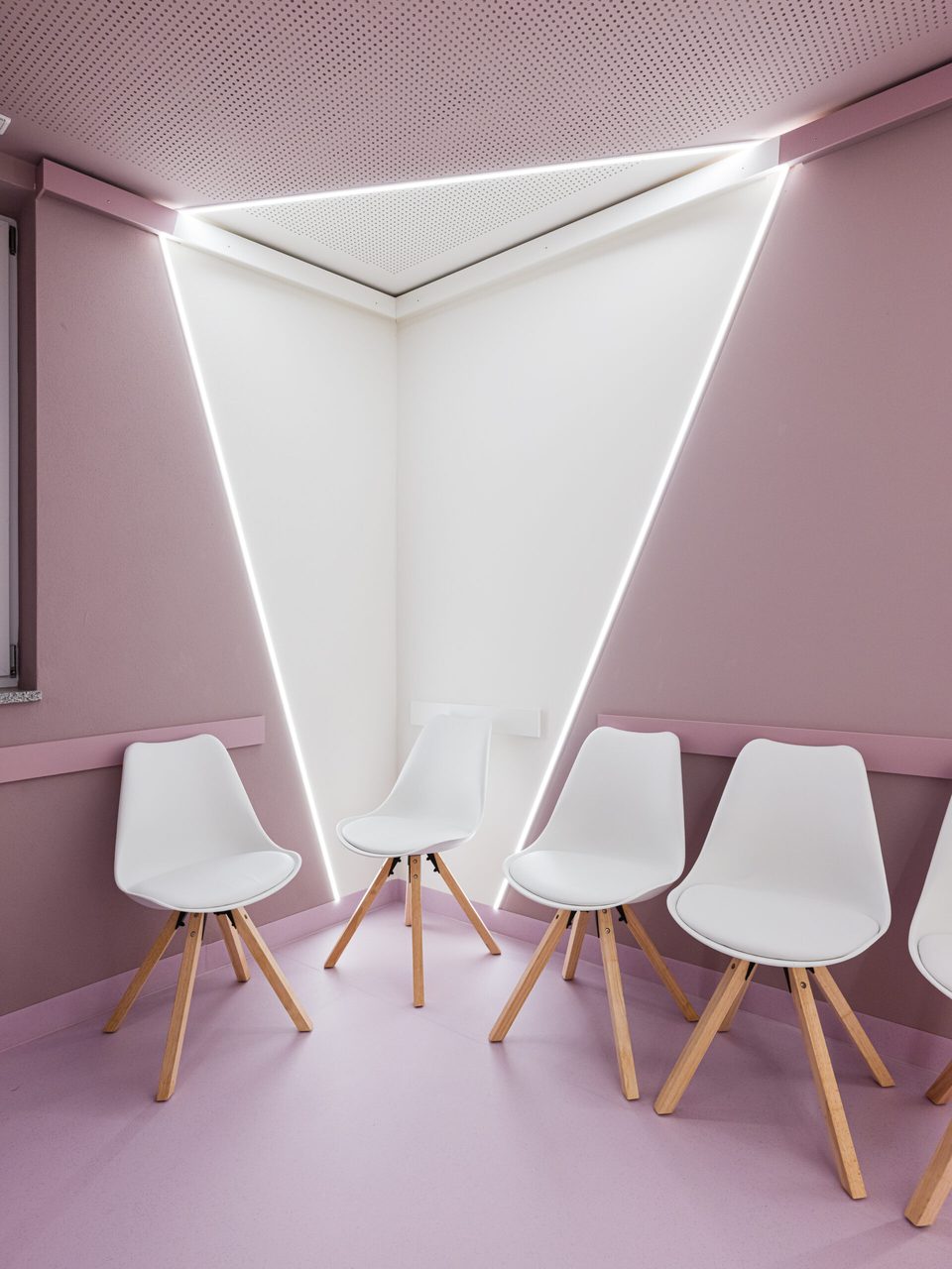 Ecke des Wartebereichs, in der ein weißer Wandkeil die roséfarbenen Flächen aufbricht, darunter die Schalenstühle