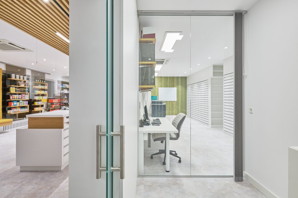Blick durch die Glastür in das Büro der Apotheke, Arbeitsplatz und Lagerregale dahinter