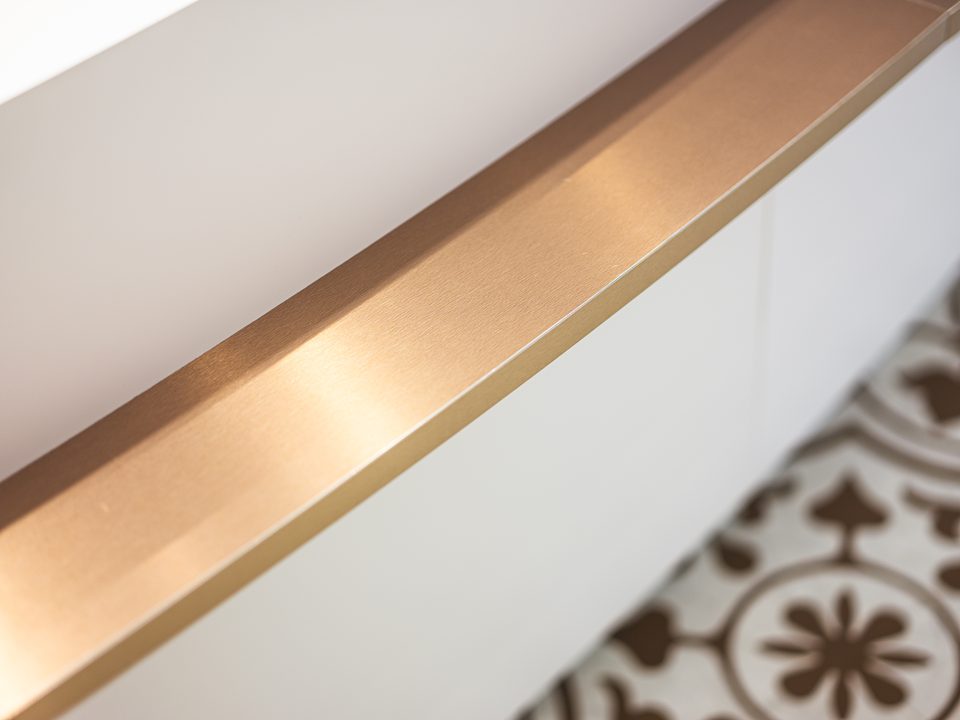 Detail einer kupferfarbenen Kante, die als schmaler Streifen durchs Bild läuft, darunter unscharf der ornamentale Fliesenboden
