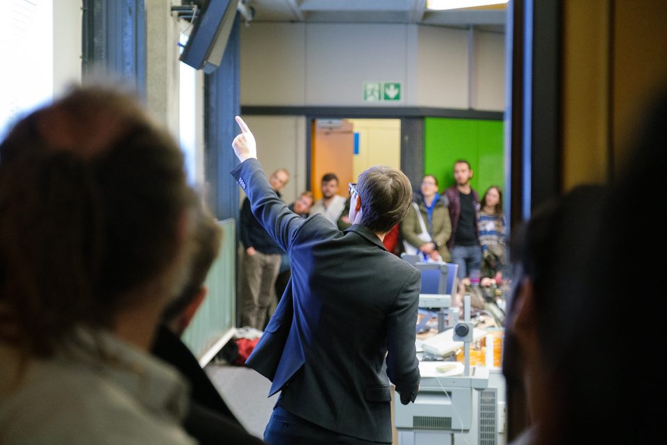 Ein Redner hebt die Hand, davor die unscharfen Köpfe des Publikums im Ausstellungsbereich