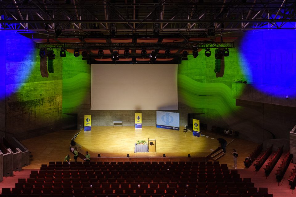 Der Audimax von oben mit blau-grün angestrahlten Betonwänden und der Leinwand über der Bühne