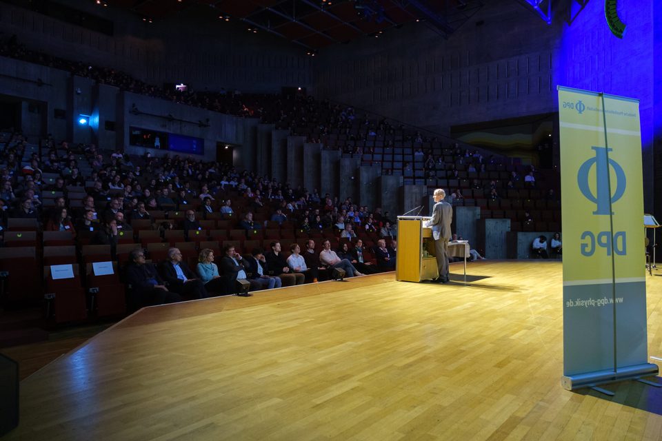 Redner am Pult im voll besetzten Audimax der Universität Regensburg, daneben die beleuchtete DPG-Stele