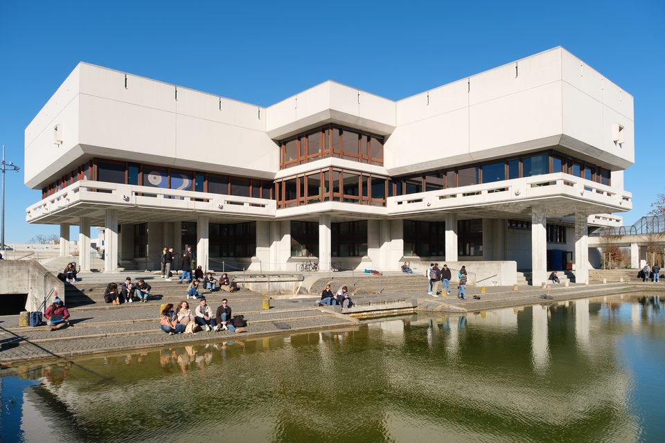 Blick über das Wasserbecken der Universität Regensburg auf den Betonbau, auf dessen Stufen Tagungsgäste in der Sonne sitzen