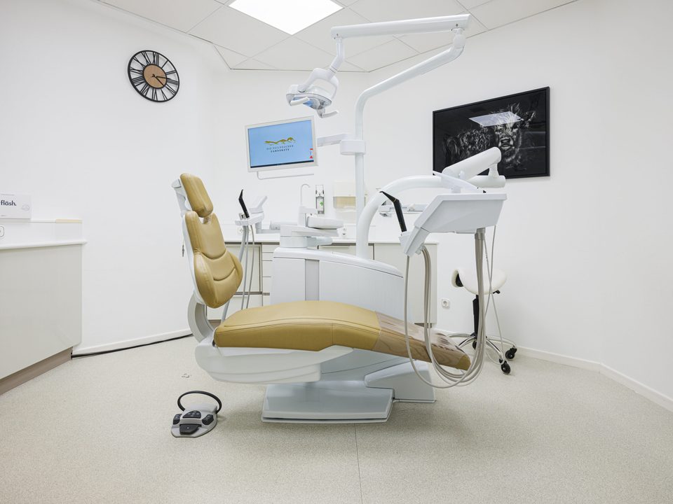 Behandlungszimmer in der Totale mit beigem Behandlungsstuhl, Monitor und OP-Leuchte vor weißen Wänden