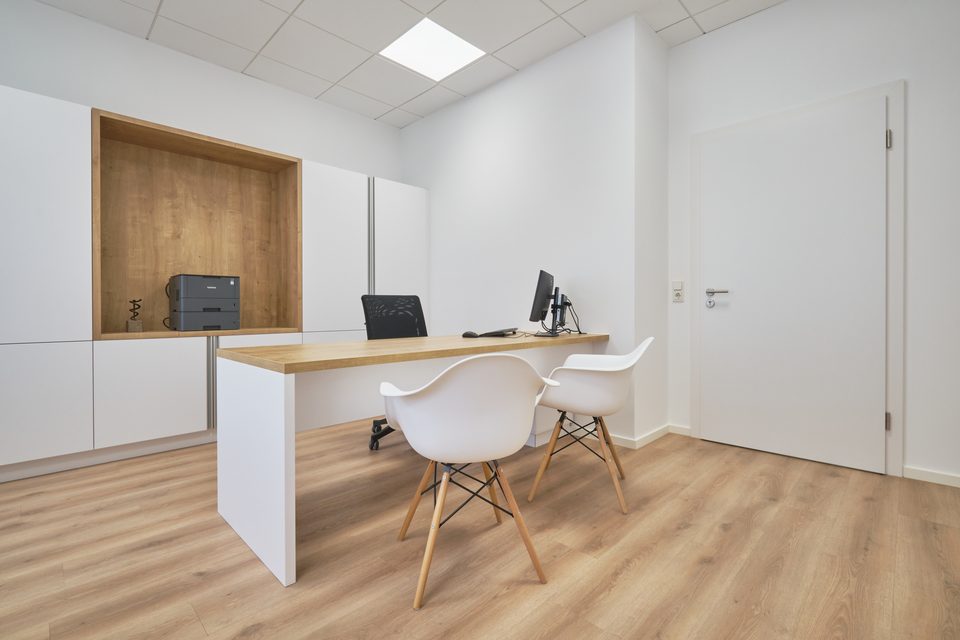 Sprechzimmer mit Schreibtisch aus hellem Holz, zwei Schalenstühlen und bündigem Einbauschrank