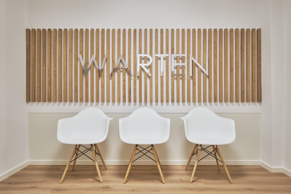 Wartebereich der Gemeinschaftspraxis Vilshofen mit dem Wort „Warten“ in Metalllettern auf der Holzlamellenwand
