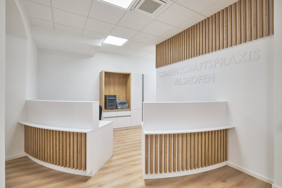 Zwei Empfangstresen mit geschwungener Holzlamellenfront, rechts an der weißen Wand die erhabenen Buchstaben Gemeinschaftspraxis Vilshofen unter einem Holzlamellenband