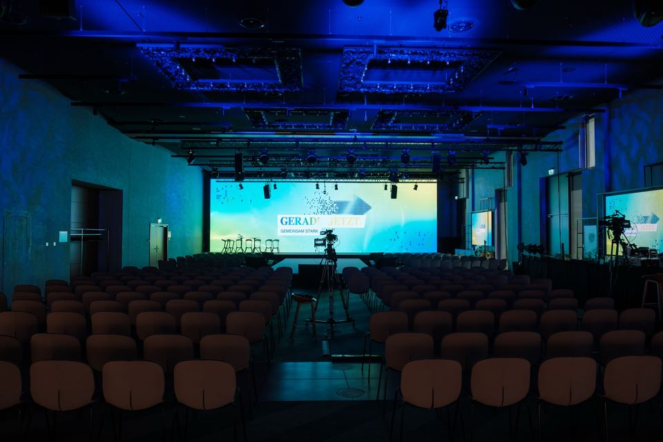 Der noch leere Saal mit Stuhlreihen in blauem Licht, vorne die beleuchtete Bühne