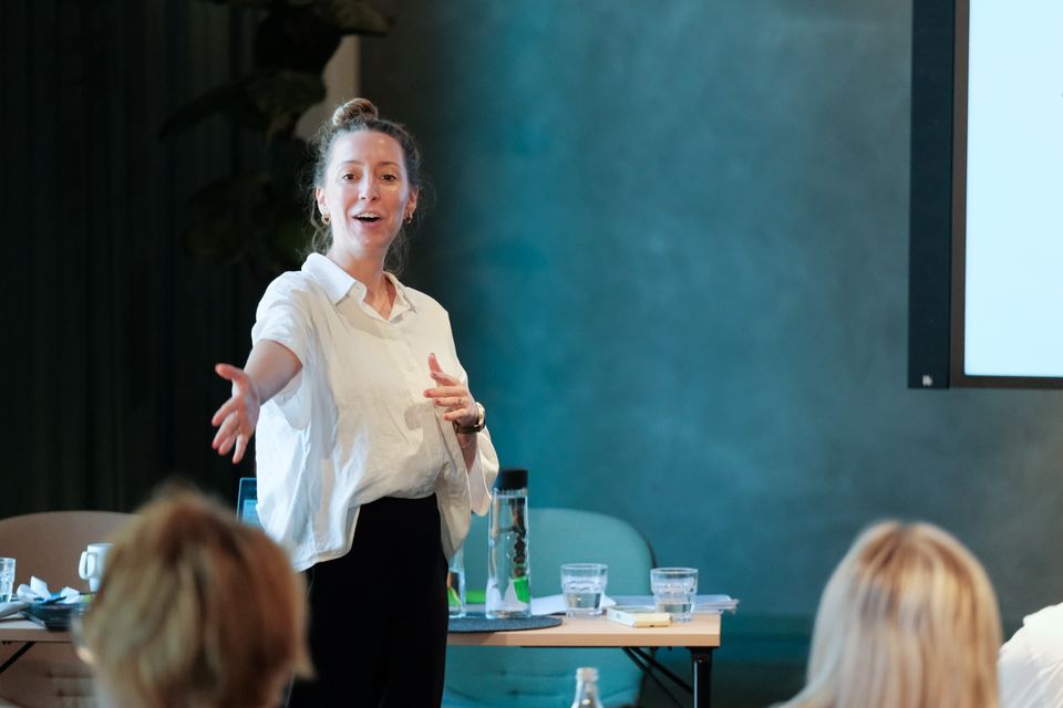 Workshopleiterin in weißer Bluse erklärt mit ausgebreiteten Armen vor der Gruppe