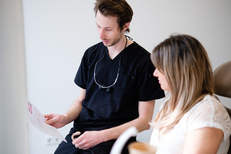 Zahnarzt in dunklem Kasack erklärt einer Patientin den Behandlungsplan und hält eine Alignerschiene