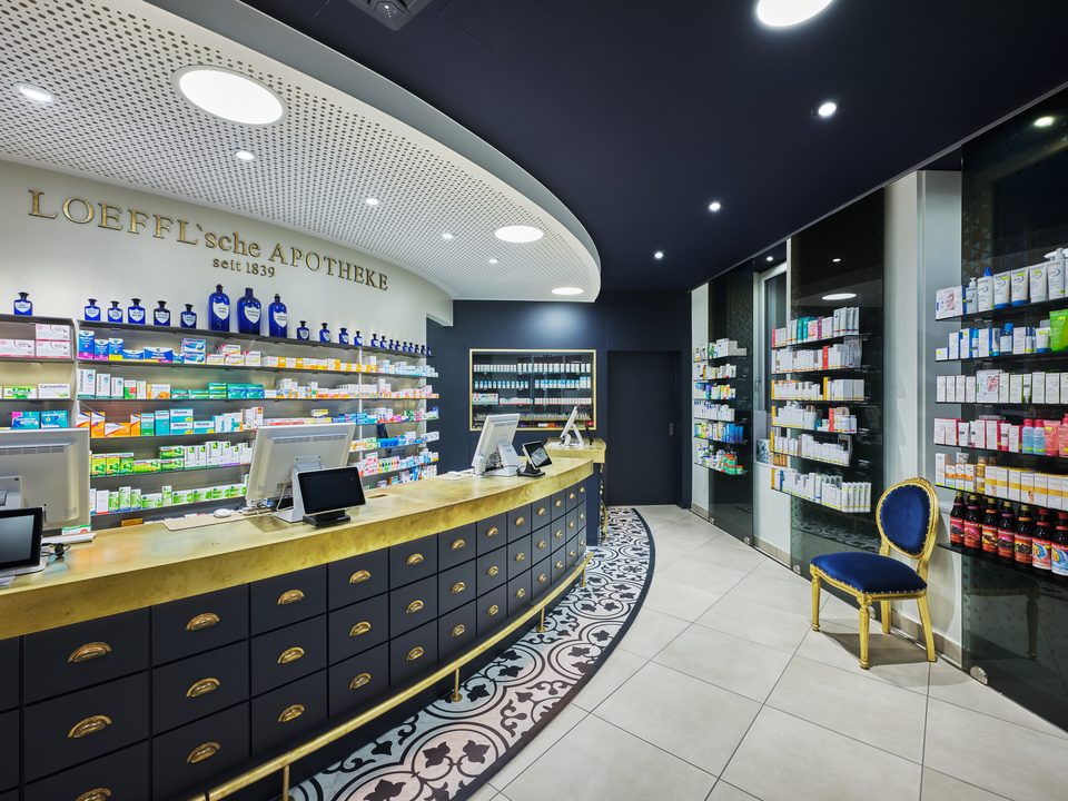 Offizin der Löffl’schen Apotheke mit geschwungenem Tresen in Navy und Messing vor der goldenen Schriftzugwand