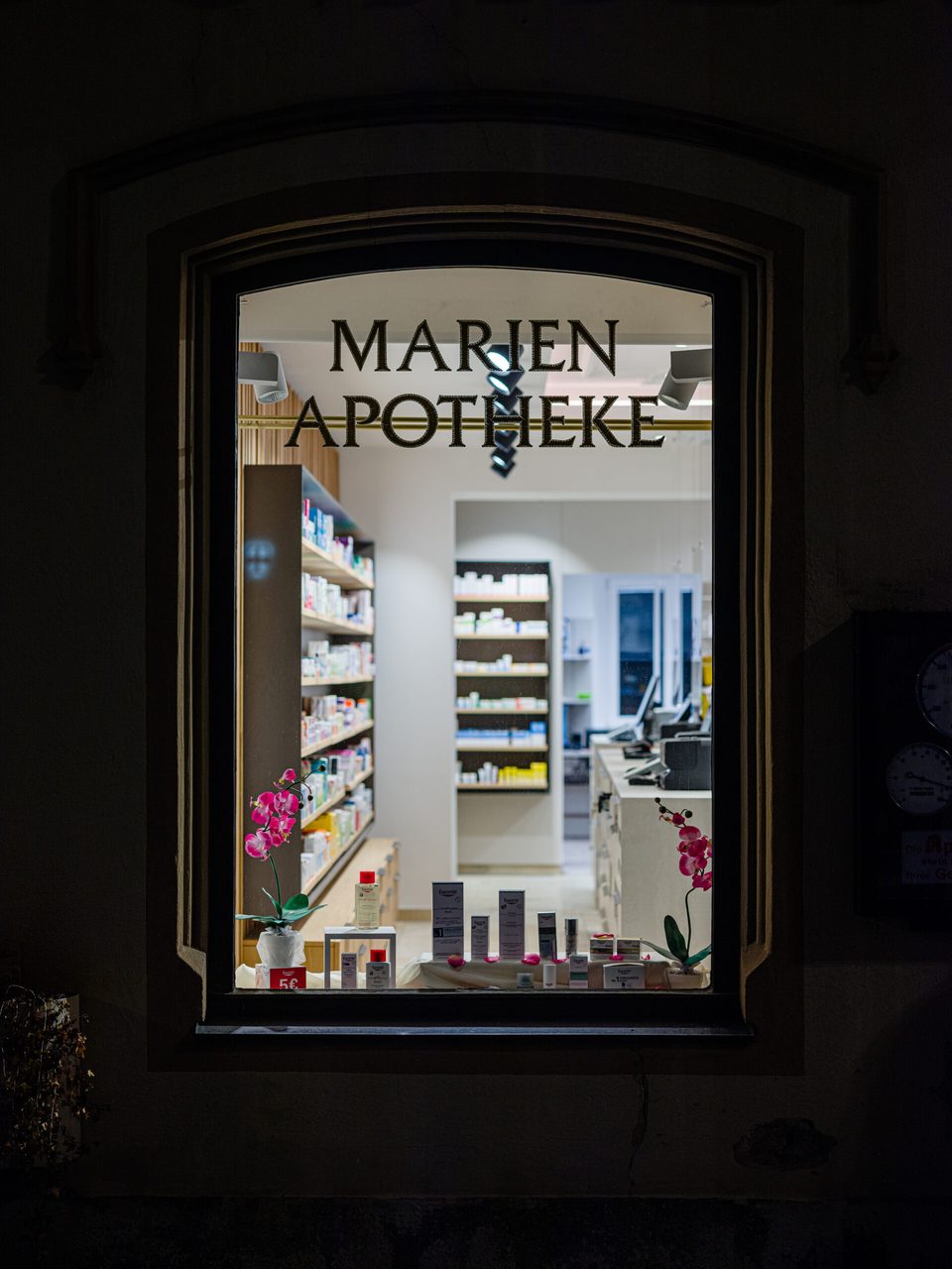 Nachtaufnahme des beleuchteten Schaufensters mit dem Schriftzug „Marien Apotheke" in der dunklen Altbaufassade