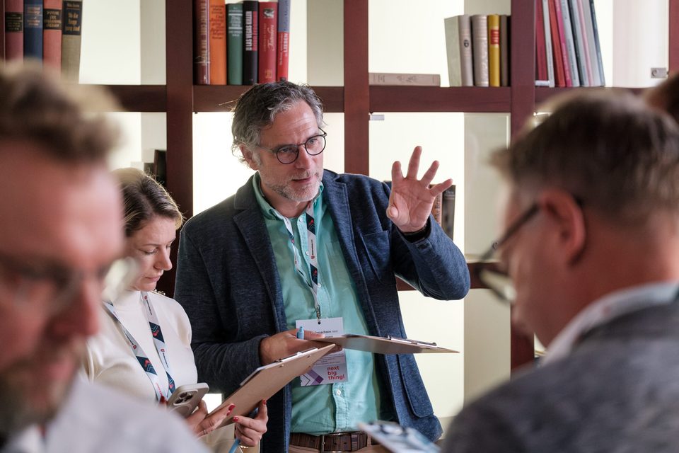 Workshop-Teilnehmer mit Klemmbrett gestikuliert vor einer Bücherwand, Zuhörende in Nahaufnahme drumherum.