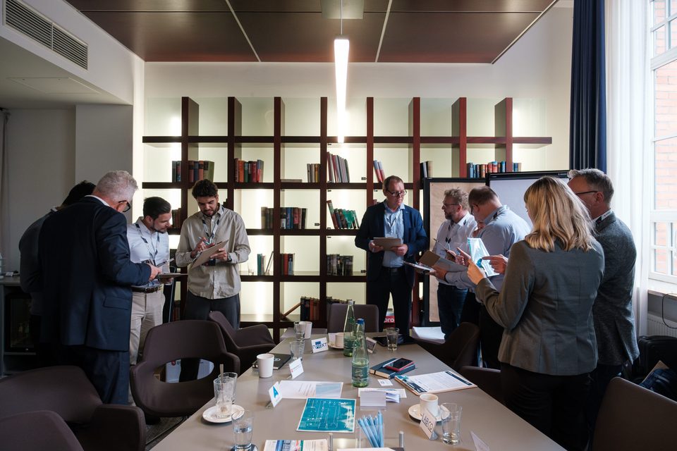 Workshop-Runde im Bibliotheksraum: Gruppen stehen im Gespräch, auf dem Tisch liegen Unterlagen und Wasserflaschen.