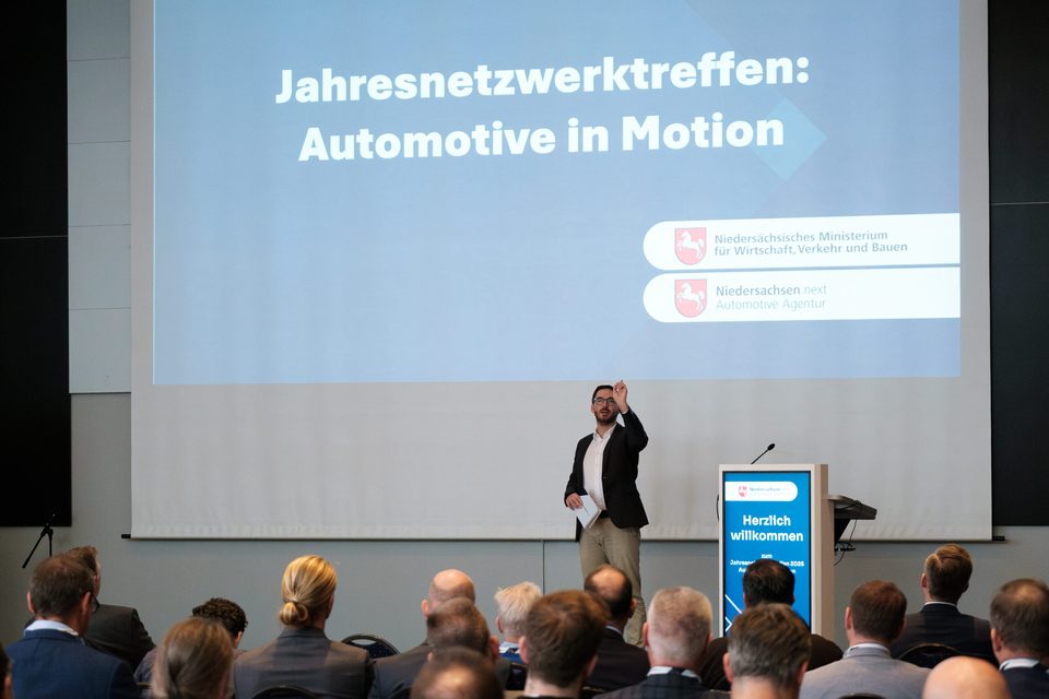 Begrüßung am Pult vor der Leinwand mit dem Leitthema „Jahresnetzwerktreffen: Automotive in Motion".