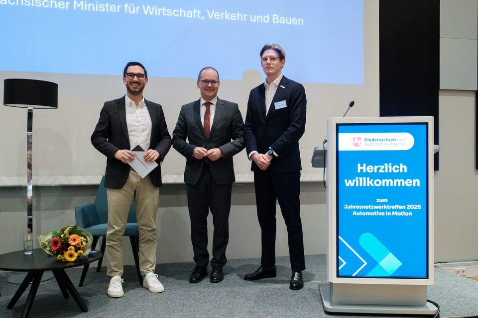 Drei Redner auf der Bühne neben der digitalen Willkommensstele zum Jahresnetzwerktreffen 2025, dahinter die Leinwand mit dem Titel des Wirtschaftsministers