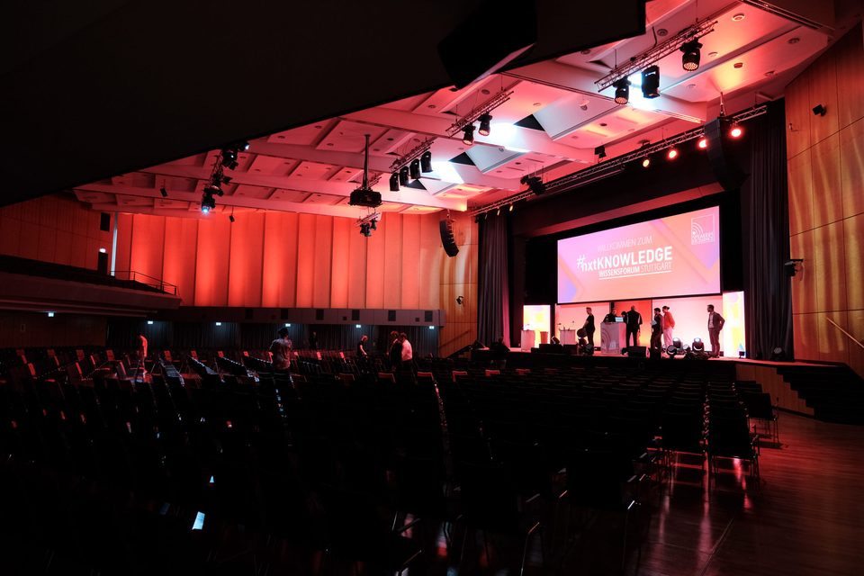 Warm rot ausgeleuchteter Saal der Kongresshalle Böblingen vor Veranstaltungsbeginn, Bühne mit #nxtKNOWLEDGE Leinwand.