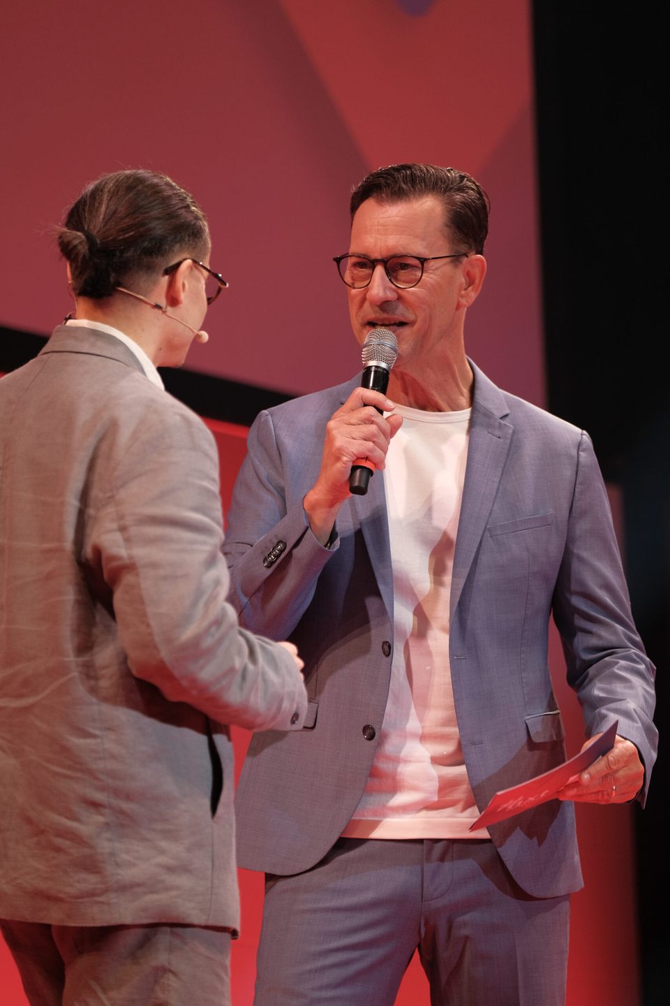 Moderator mit Mikrofon begrüßt einen Speaker auf der Bühne, beide im Gespräch vor roter Wand.