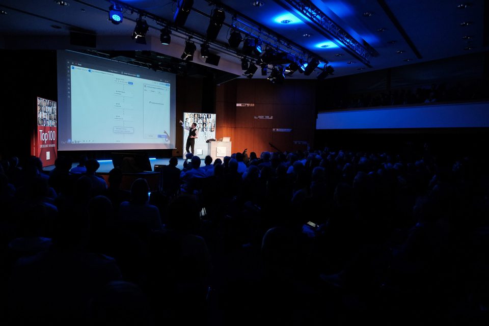 Voll besetzter Saal von hinten fotografiert, blaues Bühnenlicht und Präsentationsleinwand beim #nxtKNOWLEDGE Wissensforum.