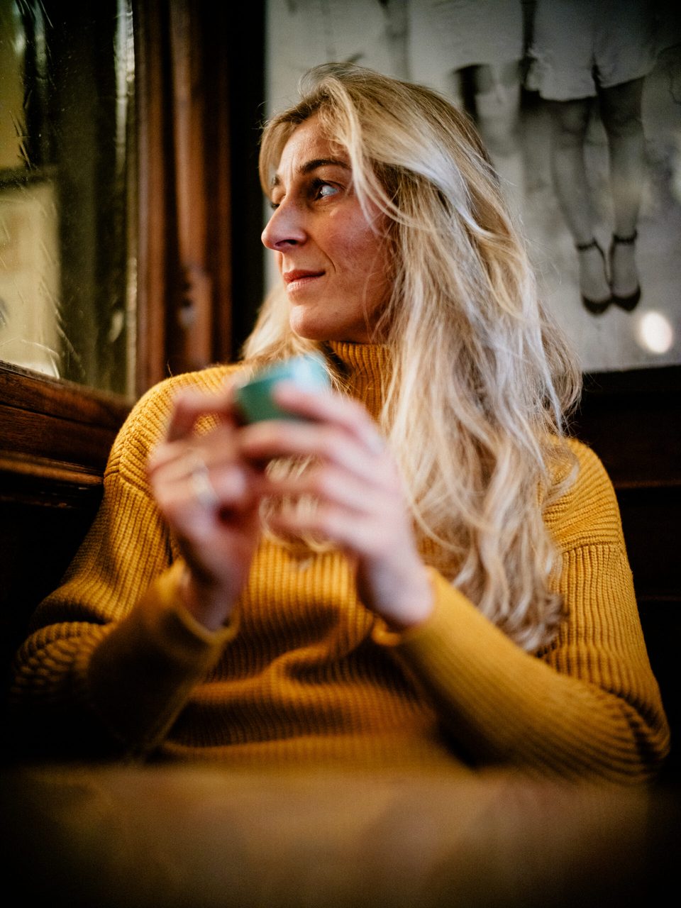 Blonde Frau mit gelbem Pullover und grüner Kaffeetasse am Tisch im Orphée