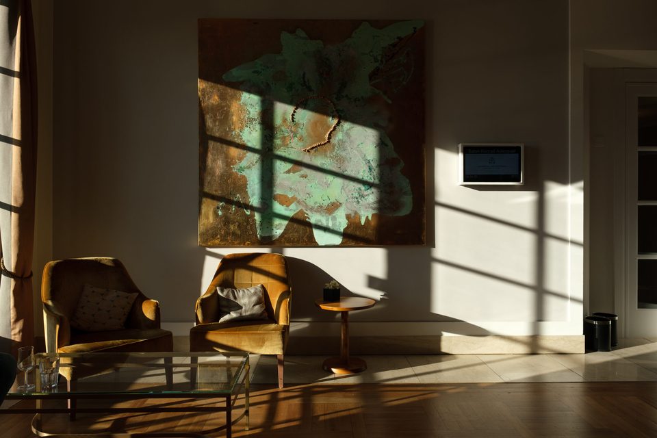 Zwei ockerfarbene Sessel vor einem großen Wandbild mit grüner Kupferpatina, quer darüber die Schatten der Fenstersprossen