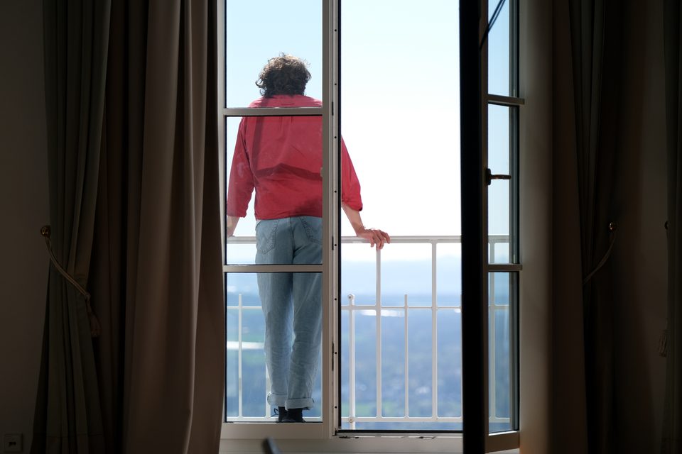 Ein Mann im roten Sweater steht auf einem Balkon mit offenen Flügeltüren und sieht in die Ferne