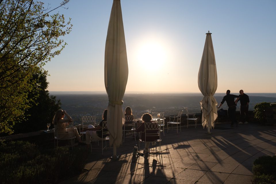 Mehrere Menschen sitzen auf der Terrasse und lehnen an der Brüstung, während sie in die tiefe Abendsonne über der weiten Landschaft sehen
