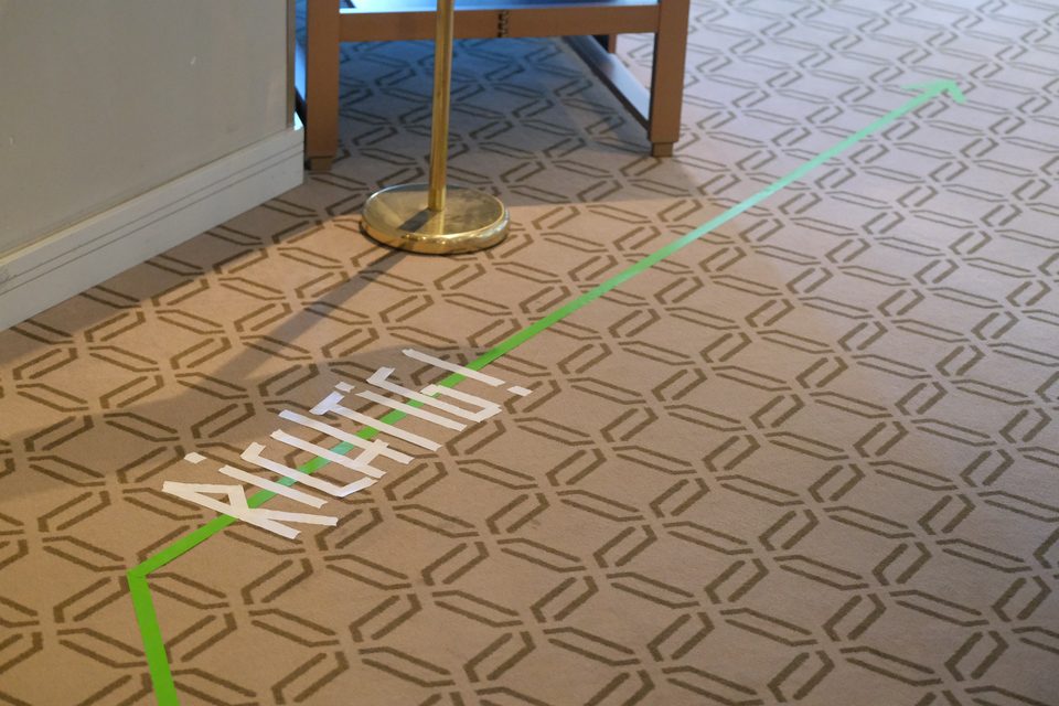 Grünes Klebeband führt als Leitlinie mit Pfeil über den gemusterten Hotelteppich, daneben das Wort „Richtig!“ in weißen Klebebuchstaben