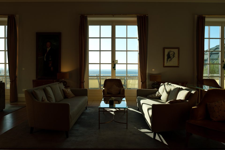 Salon mit Sofagruppe, Gemälden und bodentiefen Fenstern, durch die der Blick ins Tal geht
