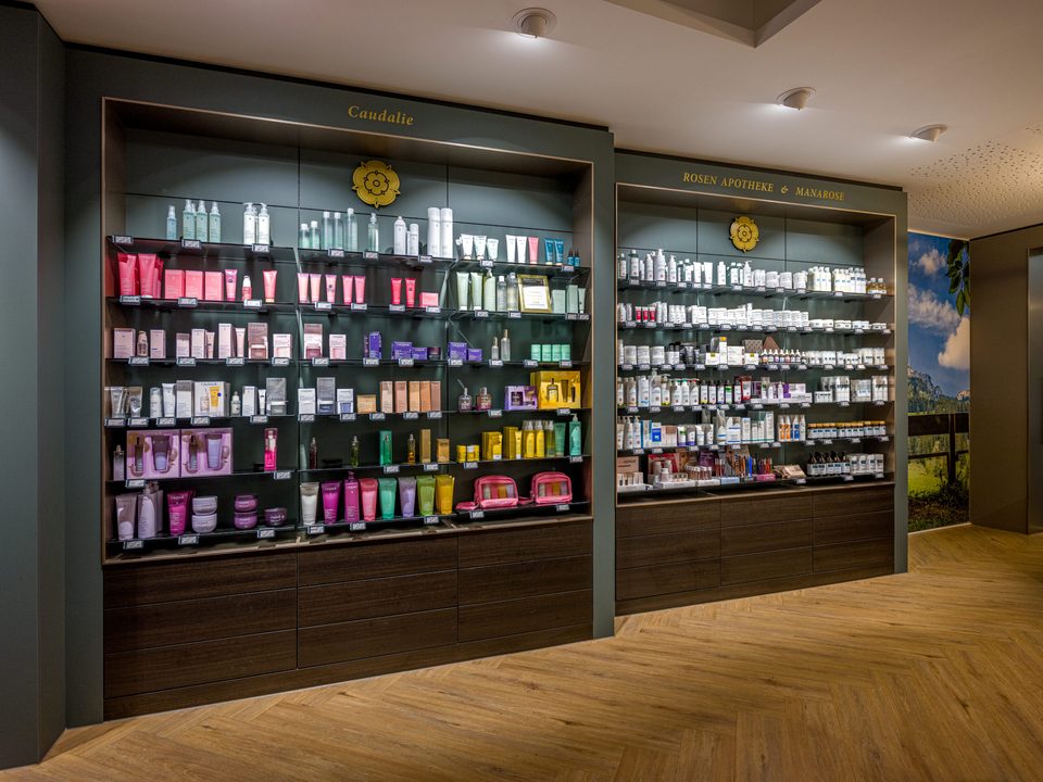 Zwei dunkelgrün gerahmte Regalfelder mit goldenen Schriftzügen „Caudalie" und „Rosen Apotheke & Manarose", gefüllt mit Kosmetik