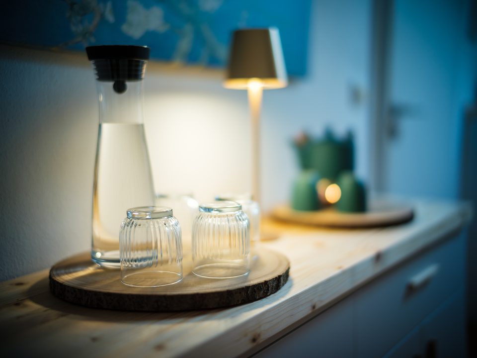 Wasserkaraffe und Gläser auf einem runden Holzbrett, im Hintergrund unscharf eine kleine Lampe