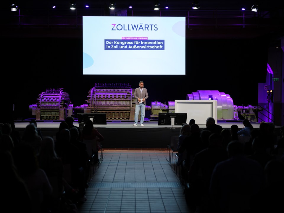 Blick über das Publikum auf die Bühne der Grand Hall, auf der Leinwand der Schriftzug Zollwärts, Der Kongress für Innovation in Zoll und Außenwirtschaft