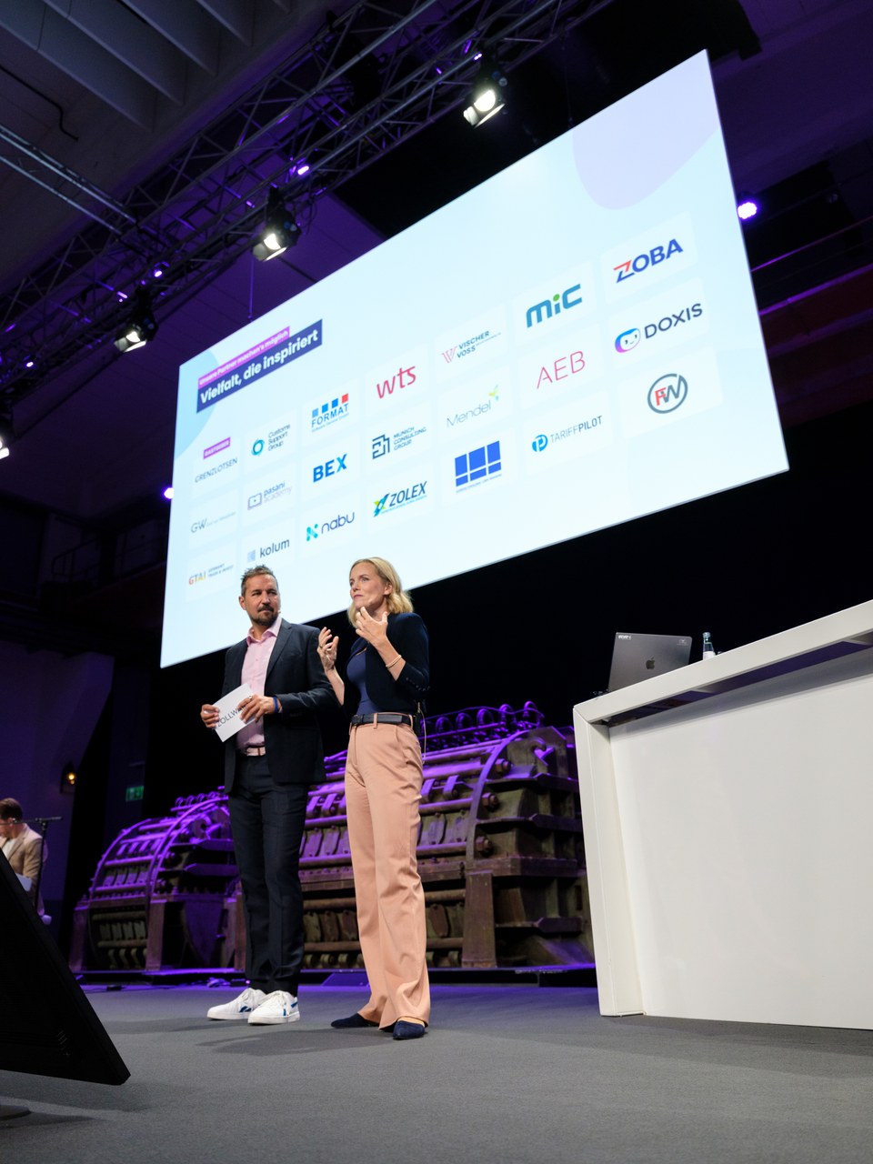 Moderationsduo auf der Bühne vor der großen Leinwand mit den Partnerlogos