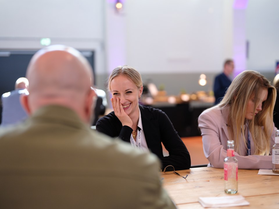 Workshop am Tisch, eine Teilnehmerin lacht, im Vordergrund unscharf ein Hinterkopf