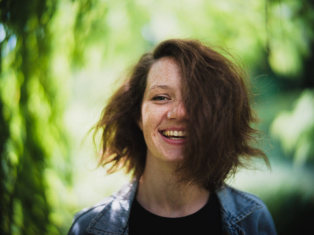 Kopfportrait einer lachenden, jungen Frau im diffusen Licht und viel Hintergrundunschärfe im Stadtpark Regensburg