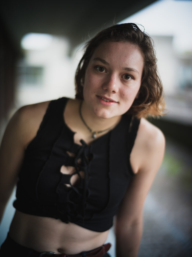 Oberkörper-Portraitfoto einer jungen Frau auf dem Gelände der Universität Regensburg, lächelnd, direkter Blick zum Betrachter.