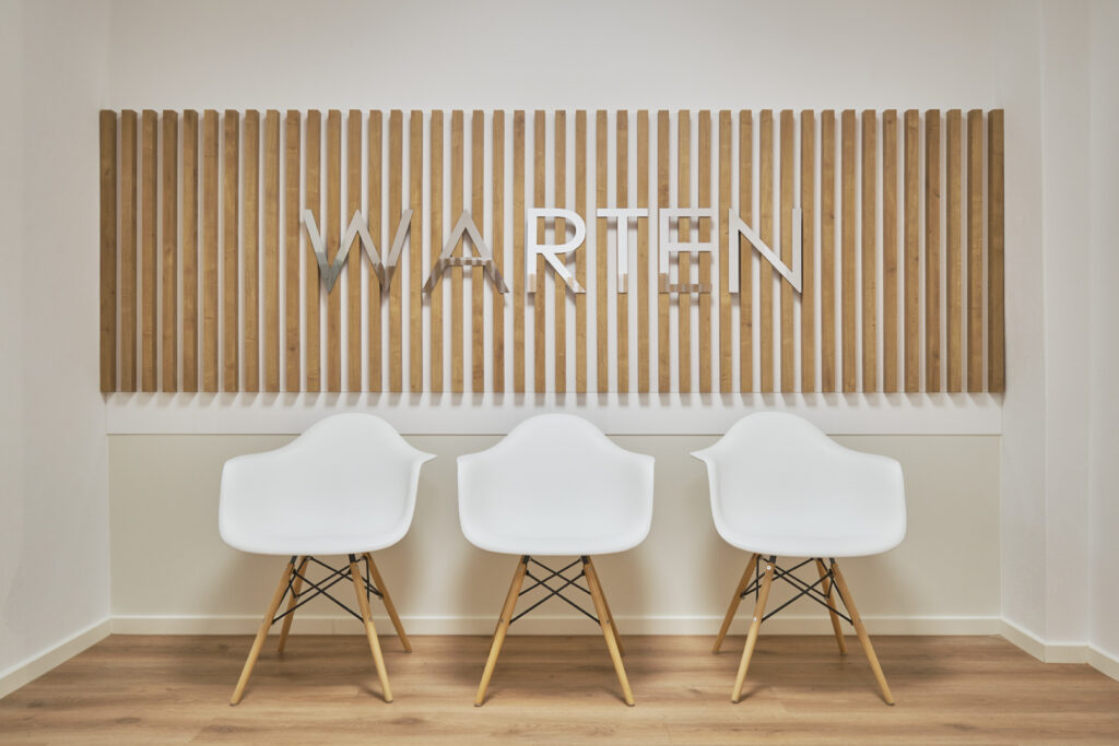 Wartebereich mit dem Wort „Warten“ in reflektierenden Buchstaben an der Wand der Gemeinschaftspraxis Vilshofen, Innenarchitektur / Interior Design von Planquelle