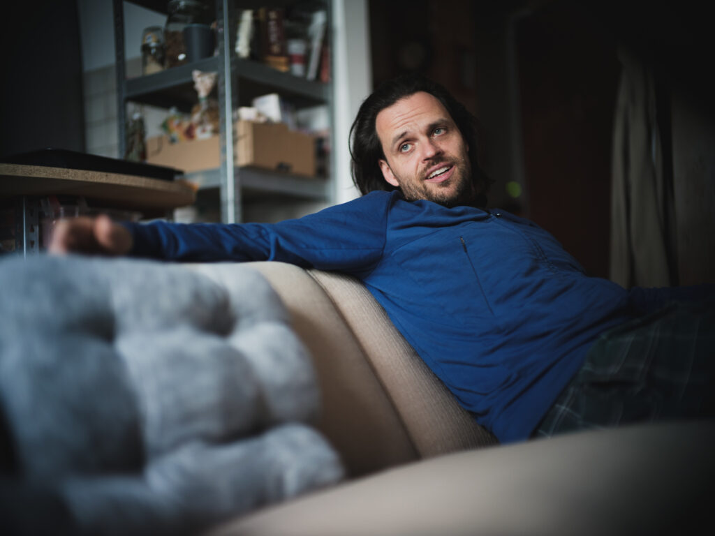 Mann mit langen, dunklen Haaren und blauen Augen lässig angelehnt im Gespräch auf einer Couch in seiner Regensburger Wohnung