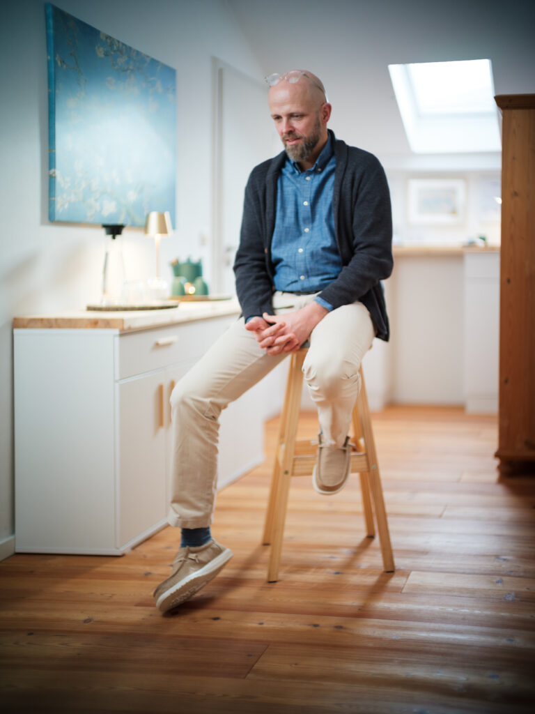 Portrait eines Mannes und Coaches mittleren Alters auf einem hohen Hocker sitzend in seiner Praxis in stimmigen gelblich-bläulichen Farben