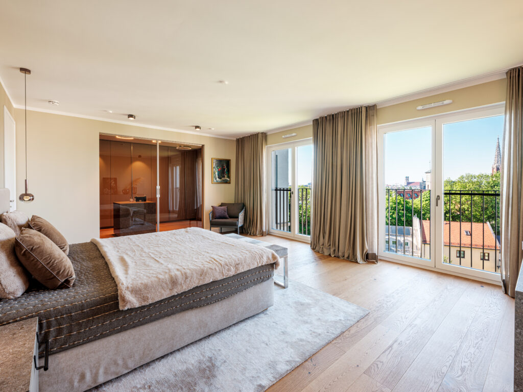 Ein modernes Schlafzimmer mit einem großen Bett, das mit einer flauschigen Decke und Kissen dekoriert ist. Es gibt eine Sitzbank am Fußende des Bettes und einen Sessel in der Ecke. Große Fenster mit beigen Vorhängen bieten einen Blick auf die Münchener Stadtlandschaft, einschließlich der Frauenkirche. Ein begehbarer Kleiderschrank ist im Hintergrund sichtbar. Die Einrichtung umfasst helle Holzböden und minimalistische Möbel.