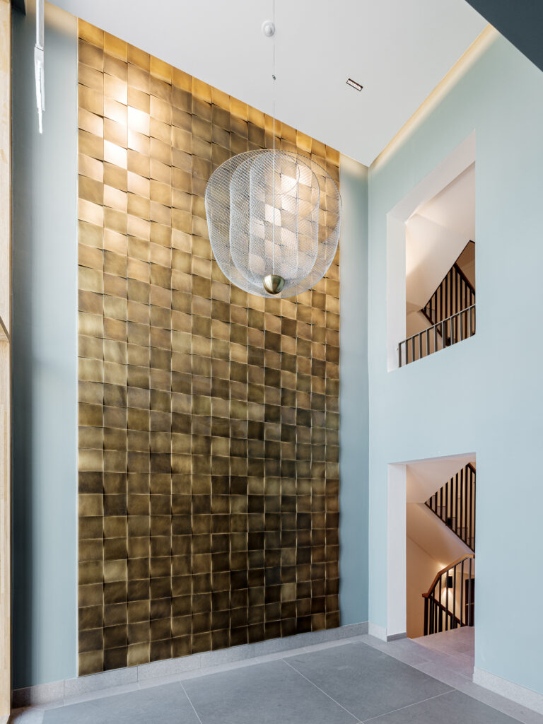 Ein moderner Eingangsbereich mit einer hohen Decke und einer auffälligen Wand aus goldenen, quadratischen Platten. Eine große, runde Pendelleuchte aus Metall hängt von der Decke. Eine elegante Treppe mit schwarzen Geländern führt in ein Obergeschoss. Der Boden besteht aus grauen Fliesen, und die Wände sind in einem hellen Blaugrün gehalten.