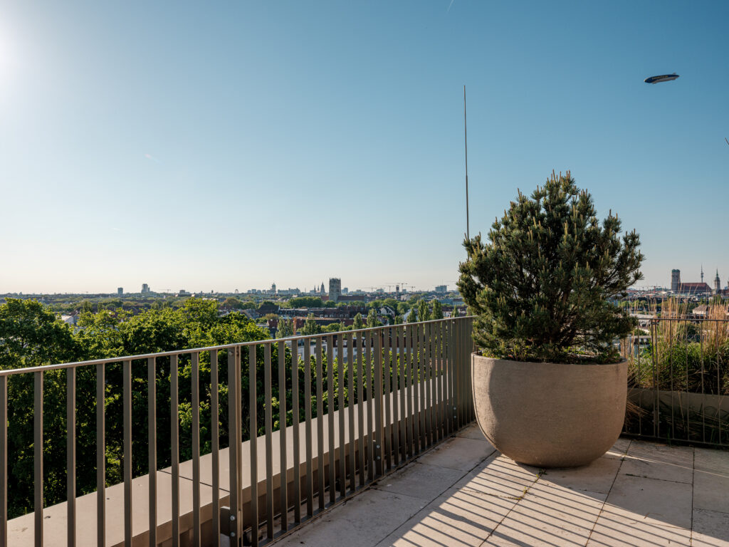 Ein Balkon mit einer Betonbalustrade und einer großen Topfpflanze. Der Blick erstreckt sich über München mit vielen Wohnhäusern, Bäumen und einem Kirchturm in der Ferne. Ein Zeppelin schwebt am klaren blauen Himmel, und der Balkon hat eine minimalistische Gestaltung mit hellen Bodenfliesen.