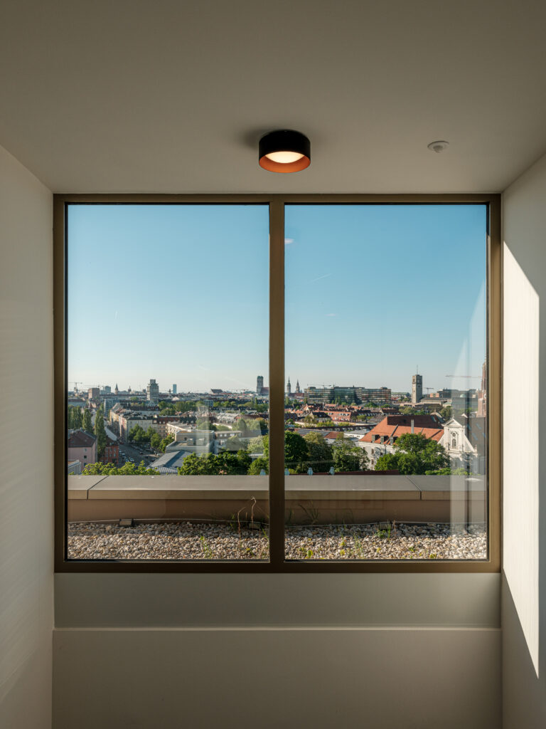Ein großes Fenster mit einem Blick auf München, einschließlich moderner Wohnhäuser, Bäume und mehrerer Kirchtürme in der Ferne. Der Himmel ist klar und blau, und der Fensterrahmen ist in einem dunklen Holzton gehalten. Kieselsteine sind entlang der Fensterbank verteilt.