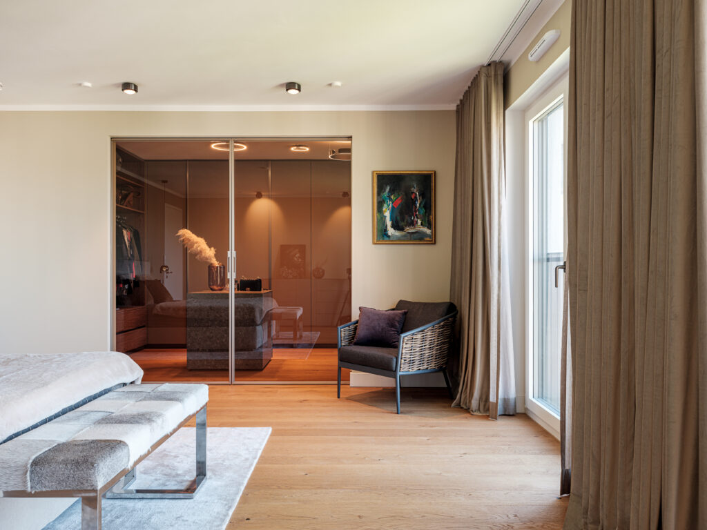 Ein stilvolles Schlafzimmer mit Holzboden und großen Fenstern, die mit beigen Vorhängen versehen sind. Im Vordergrund steht eine gepolsterte Bank mit grauem und weißem Fellbezug. Daneben befindet sich ein grauer Sessel mit einem lila Kissen. An der Wand hängt ein farbenfrohes Gemälde. Im Hintergrund führt eine Glastür zu einem Ankleidezimmer mit warmem Licht und modernen Schränken, in denen Kleidung sichtbar ist. Die Decke ist mit mehreren Einbauleuchten ausgestattet.