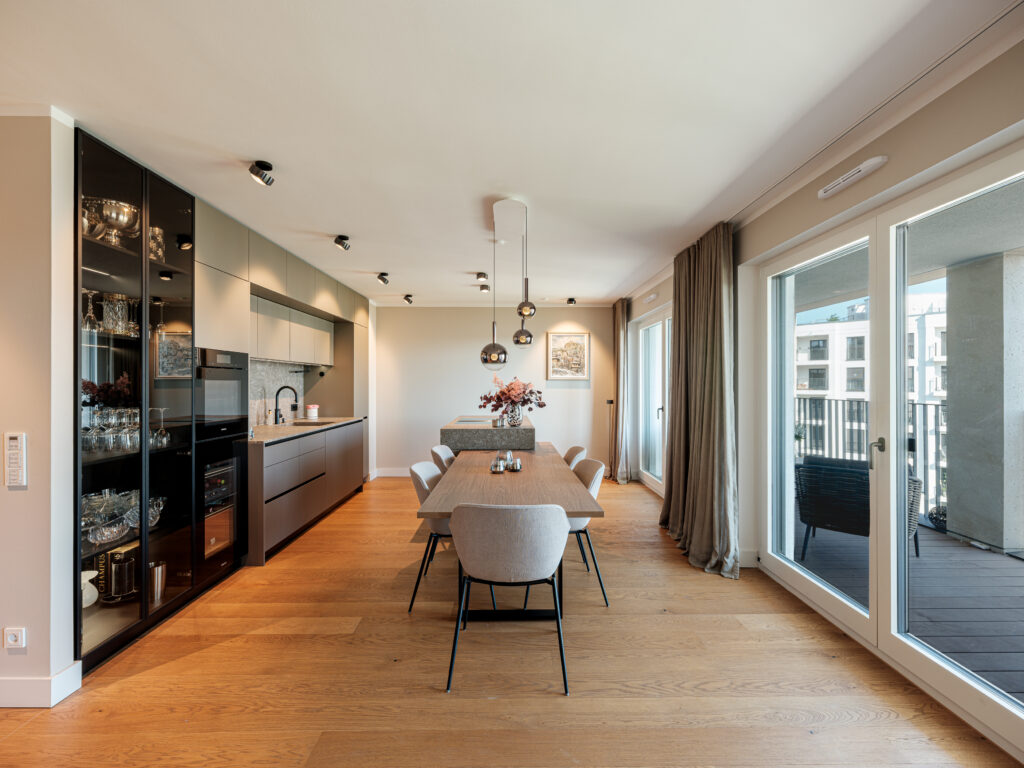 Ein offener Wohnbereich mit einer modernen Küche und einem Essbereich. Die Küche hat graue Schränke, eine graue Arbeitsplatte und eine Spüle mit schwarzem Wasserhahn. Ein großer Esstisch aus Holz mit sechs grauen Stühlen steht vor der Küche. Über dem Tisch hängen zwei runde, moderne Pendelleuchten. Große Fenster mit beigen Vorhängen führen zu einem Balkon mit Blick auf eine Stadtlandschaft.