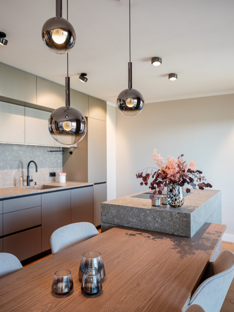 Ein moderner Essbereich mit einer offenen Küche. Über einem großen Holztisch hängen drei runde, reflektierende Pendelleuchten. Auf dem Tisch steht eine kleine, getönte Glasvase. Im Hintergrund ist eine Küchenzeile mit grauen Schränken, einer Spüle und einem Wasserhahn zu sehen. Auf der Arbeitsplatte aus Stein steht eine hohe Vase mit einem üppigen Arrangement aus rosa und braunen Trockenblumen. Die Decke hat mehrere Einbauleuchten, und die Wände sind in neutralen Tönen gehalten.