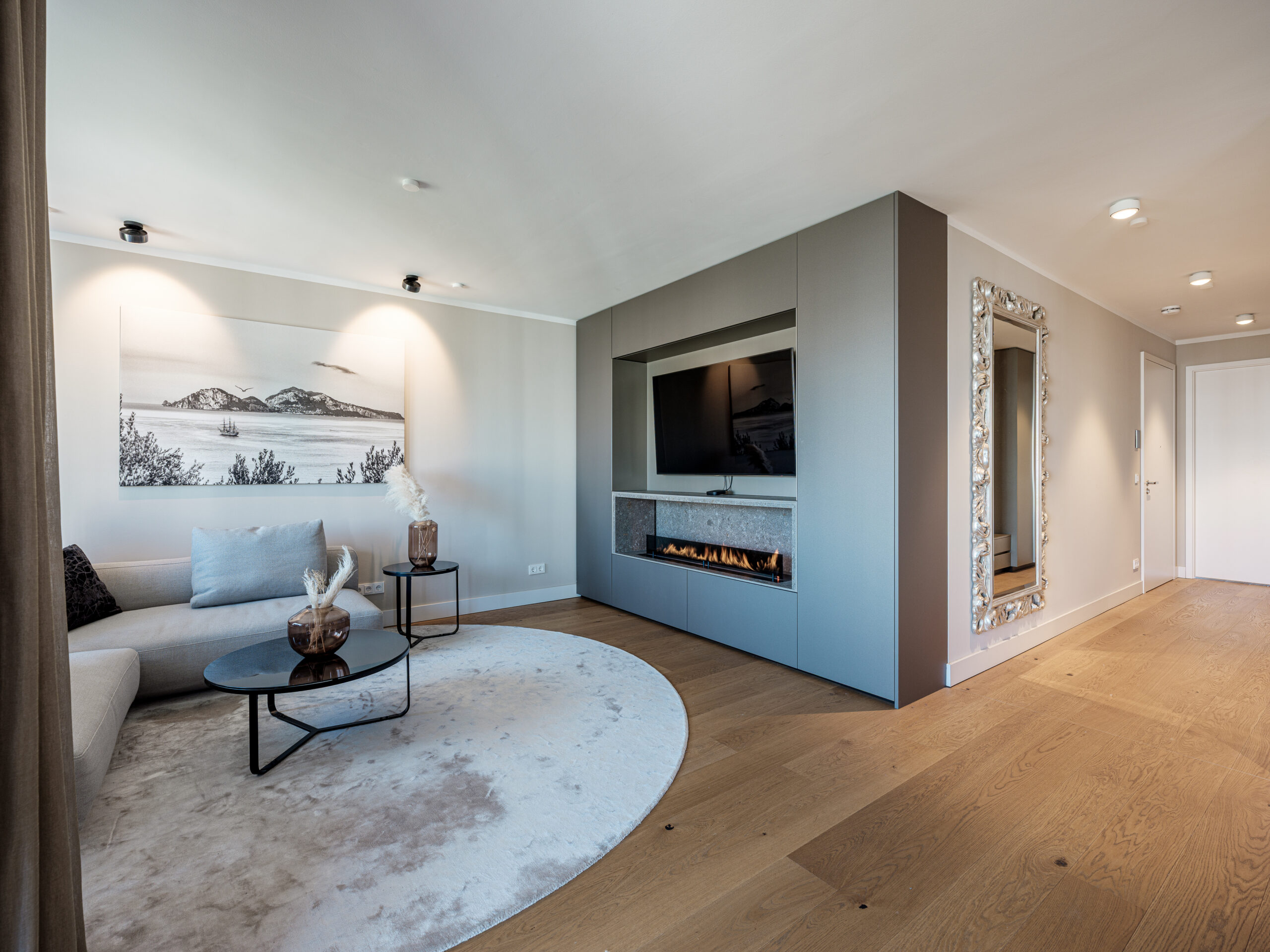 Ein stilvolles Wohnzimmer mit einem grauen Sofa, das mit Kissen dekoriert ist, und einem runden Couchtisch aus Glas. Eine moderne Feuerstelle ist in eine graue Wand mit einem Fernseher darüber eingebaut. Ein großes Schwarz-Weiß-Kunstwerk hängt an der Wand, und zwei braune Vasen mit getrockneten Pflanzen stehen auf einem Beistelltisch. Ein verschnörkelter Spiegel hängt an der Wand, und ein Teppich liegt auf dem hellen Holzboden.