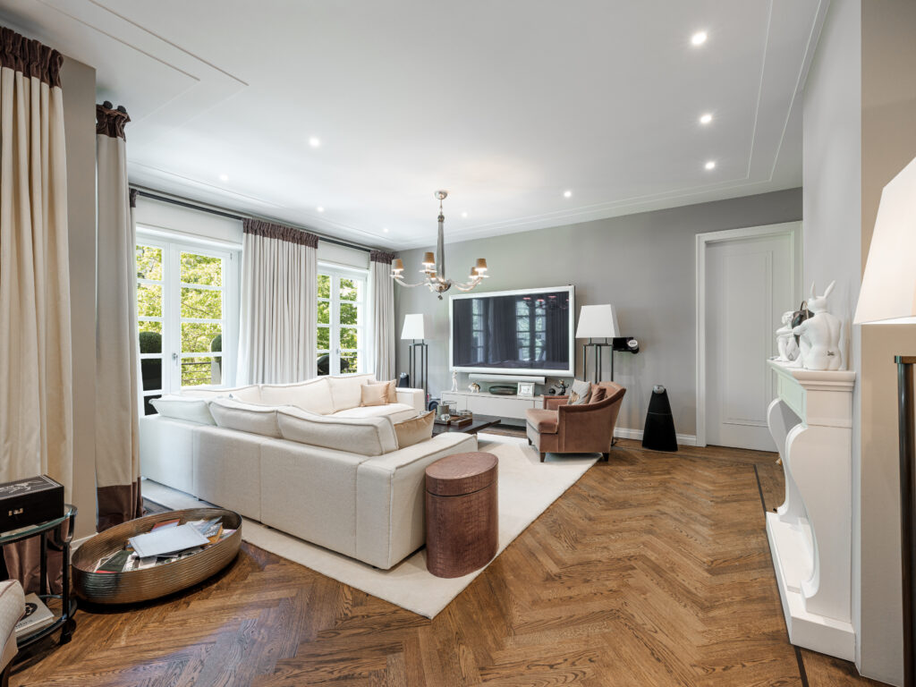 Ein stilvolles Wohnzimmer mit einem weißen Sofa, einem braunen Sessel und einem großen Fernseher an der Wand. Über einem Couchtisch hängt ein Kronleuchter, und neben dem Fernseher stehen zwei Stehlampen. Ein weißer Kamin mit Dekofiguren ist an der rechten Wand, und große Fenster mit beigen Vorhängen bieten Ausblick nach draußen. Der Boden ist mit Parkett und einem hellen Teppich ausgelegt.